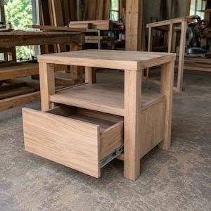 Peut inclure: Une table de chevet en bois brun clair dotée d'un plateau carré, d'une étagère ouverte et d'un tiroir coulissant avec des glissières métalliques latérales visibles. Le meuble est en bois naturel avec un grain apparent et une finition mate.