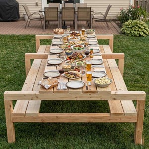 May include: A large, unfinished wooden picnic table set for a meal outdoors. The table is laden with plates of food, glasses, and bottles. The table is surrounded by green grass and other outdoor furniture.