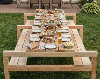 Può includere: Un grande tavolo da picnic in legno non trattato apparecchiato per un pasto all'aperto. Il tavolo è carico di piatti di cibo, bicchieri e bottiglie. Il tavolo è circondato da erba verde e altri mobili da esterno.