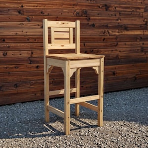 May include: A tall, unfinished wooden bar stool with a slatted back and seat. The stool has four legs connected by horizontal supports and decorative brackets. The wood has a natural, light color, and the stool is set against a wooden wall.
