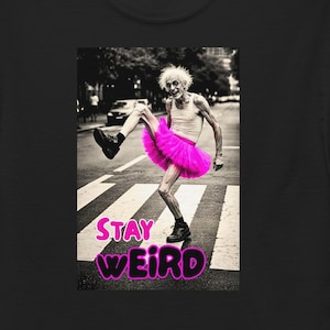 Op de afbeelding: Een zwart t-shirt met een grafische print van een ouder persoon met wild wit haar, gekleed in een felroze tutu en een wit hemdje, dansend op een zebrapad. De tekst "STAY WEIRD" staat eronder in dikke, roze, bubbelachtige letters gedrukt.
