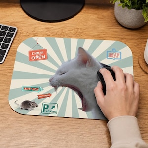 Op de afbeelding: Een rechthoekige muismat met een lichtblauwe en crèmekleurige zonnestraalachtergrond, voorzien van een grote gapende witte kat, een kleine grijze muis en diverse bordjes met de tekst Come in we're OPEN, THIS WAY, BEST BURGERS, 24/7 en P PARKING.