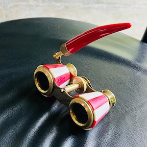 Op de afbeelding: Vintage operaglas met een rode handgreep en rood-wit gestreepte cilinders. De verrekijker heeft goudkleurige metalen accenten. De handgreep is gebogen en bevestigd aan de bovenkant van de verrekijker.