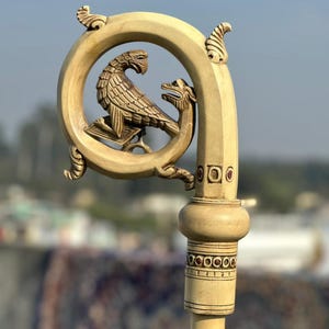Pode incluir: Um bastão de madeira esculpida com um topo curvo. O topo apresenta uma escultura detalhada de um pássaro e um dragão dentro de uma moldura circular. O bastão tem faixas decorativas e um acabamento liso e de cor clara, mostrando um artesanato intrincado.