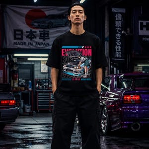 May include: A black short-sleeve t-shirt featuring a graphic print of a person leaning over the open hood of a sports car. The design includes the text "EVOLUTION" in bold pink letters, "Authentic" in script, and Japanese characters.