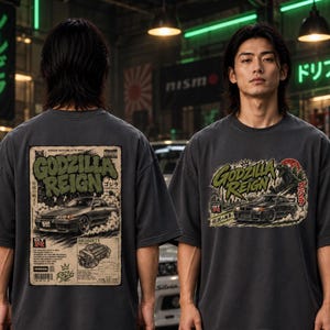 Pode incluir: Uma t-shirt cinzento-carvão com uma impressão gráfica de um Nissan Skyline GT-R R32. O design das costas apresenta o texto Godzilla Reign, uma ilustração de carro e detalhes técnicos do motor. A frente mostra o mesmo texto e o carro.