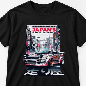 May include: A black t-shirt featuring a graphic print of a white sports car with pop-up headlights on a city street. The text reads "JAPAN'S STREET WARRIOR" and "走り屋" in large characters. The design includes red and pink accents.