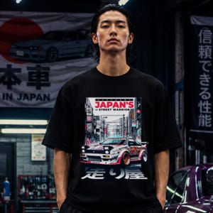 Könnte beinhalten: Ein schwarzes T-Shirt mit kurzen Ärmeln und einem Grafikdruck eines modifizierten Sportwagens auf einer Stadtstraße. Das Design enthält den Text 'JAPAN'S STREET WARRIOR' in Rot und Weiß sowie japanische Schriftzeichen über und unter dem Auto.