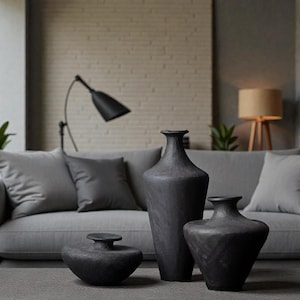 Puede incluir: Tres jarrones decorativos negros de diferentes tamaños. Los jarrones tienen superficies texturizadas y están colocados sobre una alfombra gris frente a un sofá gris. Una lámpara negra y una lámpara beige son visibles en el fondo.