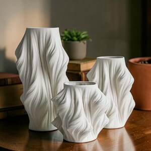 May include: Three white ceramic vases with a draped fabric design. The vases vary in size, with the tallest one on the left. The design features flowing, textured folds, creating a unique and artistic look. The vases are displayed on a wooden surface.