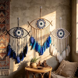 Op de afbeelding: Drie wandhangende droomvangers in de vorm van ogen, met centrale gehaakte mandala's in blauw, wit en zwart. De linker heeft zwarte veren, de middelste blauwe veren en de rechter witte veren, allemaal voorzien van kralen.