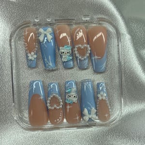 Puede incluir: Un juego de diez uñas postizas en una caja de plástico transparente. Las uñas presentan una combinación de colores azul claro y nude con varios adornos, incluyendo lazos, flores y un diseño de gato de dibujos animados. Algunas uñas tienen un diseño de punta francesa.