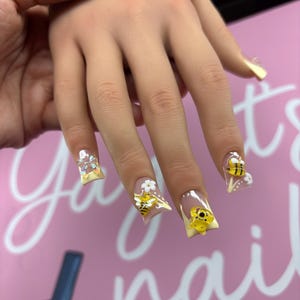 May include: Square-shaped artificial nails featuring a light yellow French tip design. The nails are decorated with three-dimensional yellow bee charms, small white flower accents, and a clear iridescent bow charm on the index fingernail.