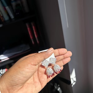 May include: Silver-coloured dangle earrings with a geometric design. The earrings feature square tops and bell-shaped bottoms adorned with small red beads. The earrings are held against a white background.