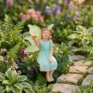 May include: A fairy figurine with light green translucent wings, wearing a light blue ruffled dress and a small floral crown. The figure has light skin, brown hair, and is holding a small yellow object in one hand while sitting upright.