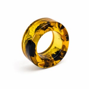 May include: A thick, circular ring made of translucent amber-colored resin with swirling black patterns throughout the material. The band has a smooth, polished finish and a wide profile, showcasing a marbled effect within the semi-transparent structure.