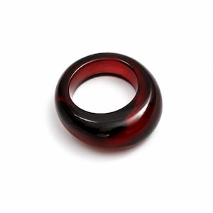 Puede incluir: Un anillo grueso y redondeado hecho de un material rojo profundo translúcido con inclusiones negras oscuras que se arremolinan por toda su superficie pulida. La banda presenta una forma circular suave y continua con un perfil ancho.