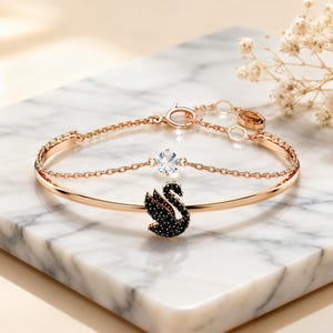 May include: A rose gold-tone bracelet featuring a swan-shaped charm encrusted with black crystals and a single clear round crystal accent. The piece includes a delicate chain, a circular clasp, and a small hexagonal tag with an engraved logo.