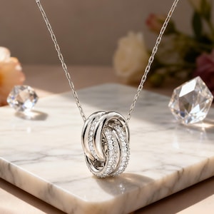 May include: A silver-toned necklace featuring a pendant composed of three interlocking circular rings. Two of the rings are smooth and polished, while the central ring is encrusted with small, clear, sparkling stones. The pendant hangs from a delicate chain.