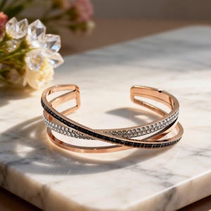May include: A rose gold cuff bracelet featuring a multi-layered design with two intersecting bands. One band is encrusted with clear white crystals, while the other is inlaid with small, square-cut black stones, creating a contrasting effect.