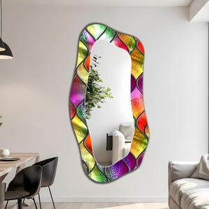 May include: A tall, irregular, wavy-edged wall mirror featuring a decorative border of multicoloured, translucent glass tiles in shades of purple, green, yellow, and orange, arranged in a repeating scalloped pattern around the central reflective surface.
