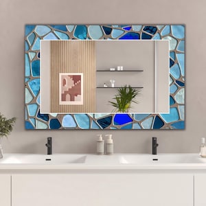 Può includere: Specchio da parete rettangolare con bordo a mosaico nei toni del blu e del grigio. Lo specchio riflette una scena di bagno con un lavabo bianco, rubinetti neri e un'opera d'arte incorniciata. Il bordo a mosaico aggiunge un tocco decorativo.