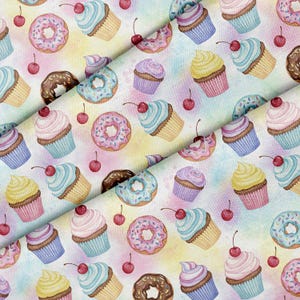 Può includere: Un motivo in tessuto con una stampa ripetuta di cupcake colorati, ciambelle glassate e piccole ciliegie rosse sparse su uno sfondo acquerellato dai toni pastello tenui con sfumature miste di giallo, blu e viola.