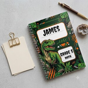 May include: A spiral-bound notebook featuring a green dinosaur illustration on a dark background with jungle leaves and orange accents. The cover displays the text "JAMES" and "GRADE 5 MATH" in white rectangular labels with orange borders.