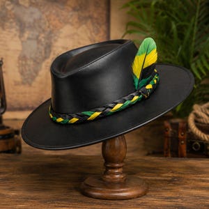 May include: A black leather hat with a wide brim and a pinched crown. The hat features a braided hatband in black, green, and yellow, accented with a single upright feather in matching green and yellow tones tucked into the side of the band.