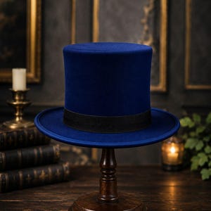 Puede incluir: Un sombrero de copa azul profundo con una textura suave similar al fieltro y una corona plana. El sombrero presenta un ala ancha y estructurada y una banda de cinta de grosgrain negra sólida que rodea la base de la corona.