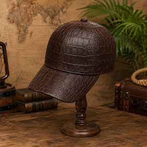 Puede incluir: Una gorra de béisbol de color marrón oscuro con un patrón texturizado de piel de cocodrilo. La gorra presenta una corona estructurada, una visera curva y un pequeño botón en la parte superior central. El material tiene un acabado brillante.
