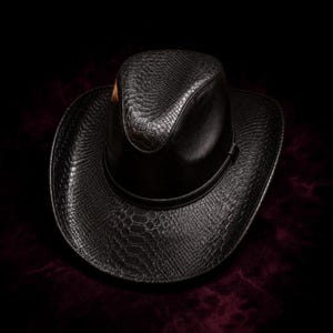 Puede incluir: Un sombrero de vaquero negro con un patrón de piel de serpiente texturizado en la copa y el ala ancha. El sombrero incluye una banda negra lisa que rodea la base de la copa, creando un contraste con la detallada textura de escamas.