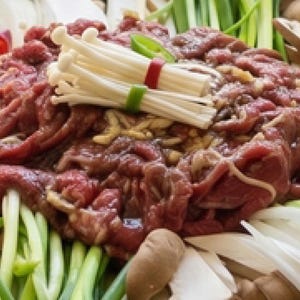 Peut inclure: Un plat de bœuf bulgogi cru composé de fines tranches de viande marinée au centre, entourées de champignons enoki blancs, de carottes orange tranchées, de champignons shiitake bruns, d'oignons verts, de quartiers d'oignons blancs et de piments rouges.