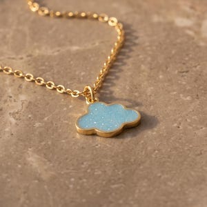 Puede incluir: Un collar de cadena en tono dorado con un pequeño colgante en forma de nube azul claro con un acabado ligeramente brillante. La delicada cadena de metal se conecta a la parte superior del dije de nube, que tiene un borde liso y superficie plana.