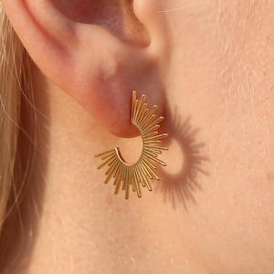 May include: A gold-toned earring shaped like a semi-circle with radiating thin metal spikes extending outward from the curved edge. The earring features a sunburst design with a polished finish, creating a distinct silhouette against the skin.