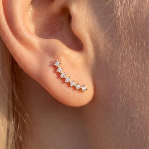 May include: A gold-toned ear climber earring featuring a curved row of seven small, clear, round-cut stones set in a line. The earring follows the natural curve of the earlobe, with each stone held in a secure metal setting, creating a delicate look.