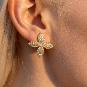 Könnte beinhalten: Ein goldfarbener Ohrstecker in Form einer vierblättrigen Blume, der vollständig mit kleinen, funkelnden klaren Kristallen besetzt ist. Der Ohrstecker befindet sich an einem Ohrläppchen und zeigt das symmetrische, spitz zulaufende Blütendesign.