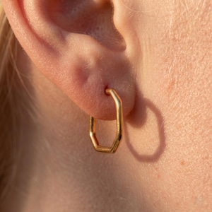 May include: A close-up of a gold-coloured, geometric hoop earring. The earring has an angular, multi-sided design, forming an octagon shape. The earring is in the ear, with the ear and skin visible in the background. The earring is a simple, modern design.