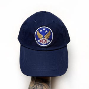 May include: Navy blue baseball cap with a circular emblem featuring gold wings, three white stars, and the letters "RPL". The cap has a curved brim and a structured crown.