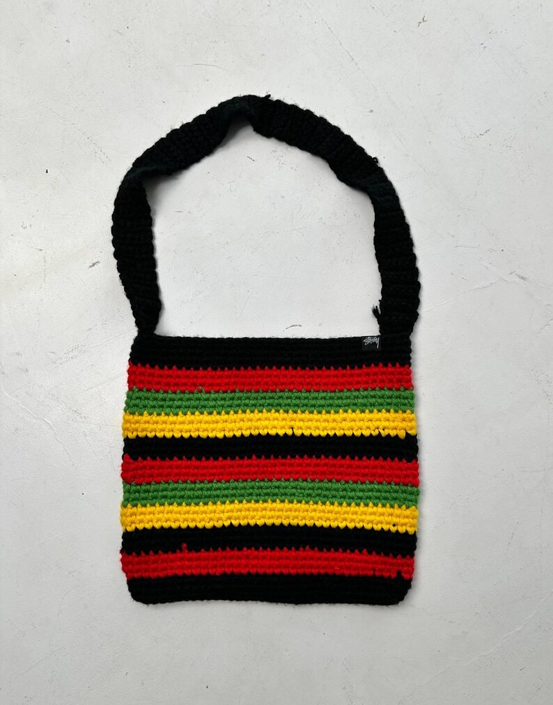 May include: A handmade crochet shoulder bag with a black strap and body. The bag features horizontal stripes in red, yellow, green, and black. The bag is made of a soft, textured yarn.