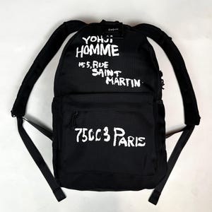 Puede incluir: Una mochila negra con texto blanco de estilo pintado a mano que dice YOHJI HOMME 155, RUE SAINT MARTIN en la parte superior y 75003 PARIS en el bolsillo delantero. La bolsa incluye correas de hombro acolchadas ajustables y un asa superior.
