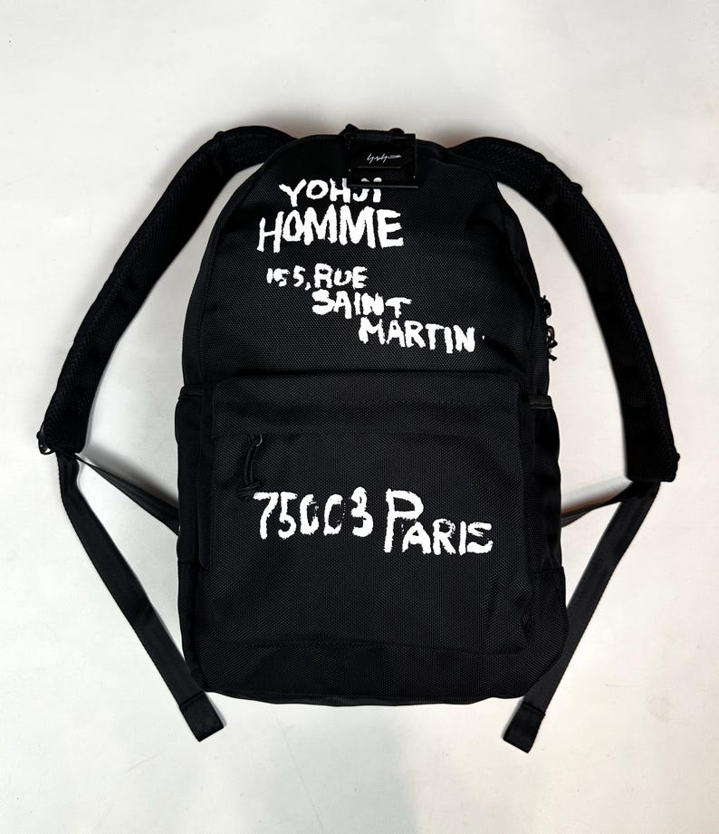 Puede incluir: Una mochila negra con texto blanco de estilo pintado a mano que dice YOHJI HOMME 155, RUE SAINT MARTIN en la parte superior y 75003 PARIS en el bolsillo delantero. La bolsa incluye correas de hombro acolchadas ajustables y un asa superior.