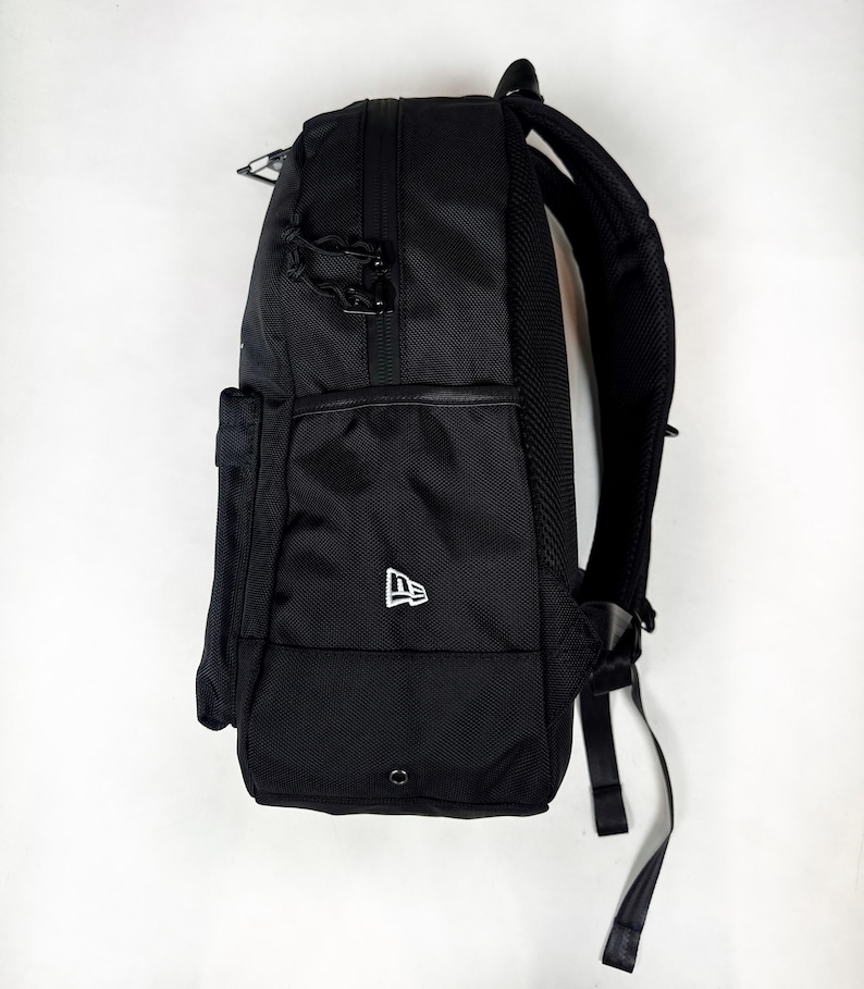 Puede incluir: Una mochila negra con exterior de tela texturizada, un bolsillo lateral y un logotipo de New Era blanco bordado en el lateral inferior. La bolsa incluye tiradores de cremallera negros, un panel trasero de malla acolchado y correas ajustables.