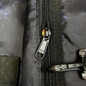 Puede incluir: Una bolsa de tela negra con cierre de cremallera YKK y tirador met&aacute;lico. El material muestra un sutil patr&oacute;n abstracto en azul oscuro y negro. Un clip de hebilla de pl&aacute;stico negro est&aacute; sujeto al lado de la cremallera en el exterior.