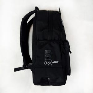 Puede incluir: Una mochila negra con correas acolchadas para los hombros y un bolsillo lateral. El lateral presenta un bloque de texto impreso en blanco que dice: Black is modest and arrogant at the same time. Black is lazy and easy - but mysterious. But above all black says this: I don't bother you - don't bother me. Debajo del texto hay una firma blanca.