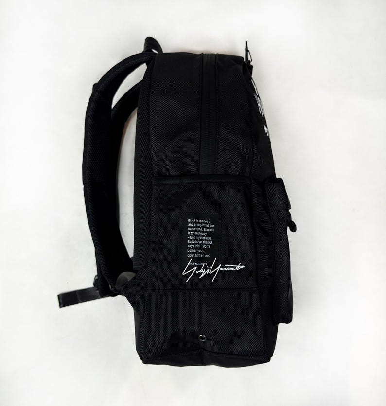 Puede incluir: Una mochila negra con correas acolchadas para los hombros y un bolsillo lateral. El lateral presenta un bloque de texto impreso en blanco que dice: Black is modest and arrogant at the same time. Black is lazy and easy - but mysterious. But above all black says this: I don't bother you - don't bother me. Debajo del texto hay una firma blanca.