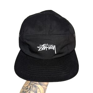 May include: Black five-panel cap with a flat brim. The cap features the white "Stüssy" logo embroidered on the front. The cap is made of a durable fabric and has a structured design.