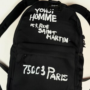 Puede incluir: Una mochila negra con texto impreso en blanco que dice YOHJI HOMME 155, RUE SAINT MARTIN 75003 PARIS. La bolsa cuenta con un compartimento principal, un bolsillo frontal con cremallera y correas ajustables, hecha de tela texturizada.