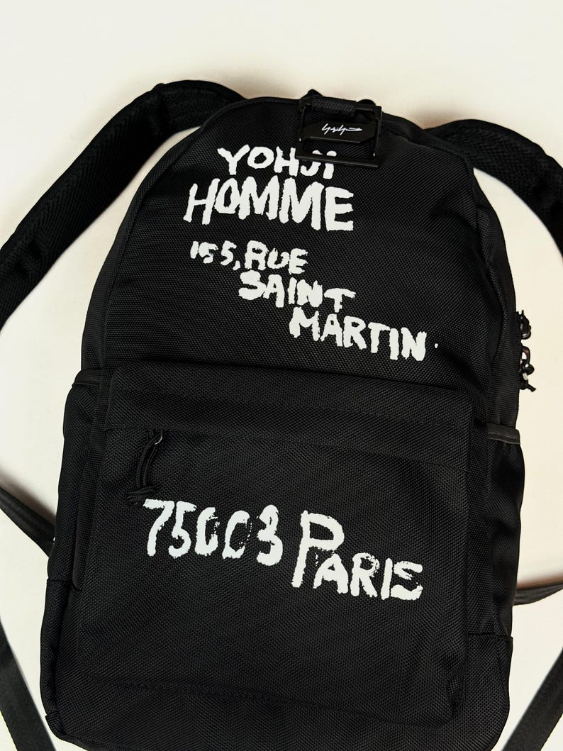 Puede incluir: Una mochila negra con texto impreso en blanco que dice YOHJI HOMME 155, RUE SAINT MARTIN 75003 PARIS. La bolsa cuenta con un compartimento principal, un bolsillo frontal con cremallera y correas ajustables, hecha de tela texturizada.
