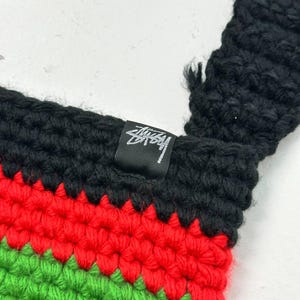 May include: Close-up of a crochet bag with a black strap and a colorful striped pattern. The stripes are red, green, and yellow, with a black top. A small black tag with white text is sewn into the bag.